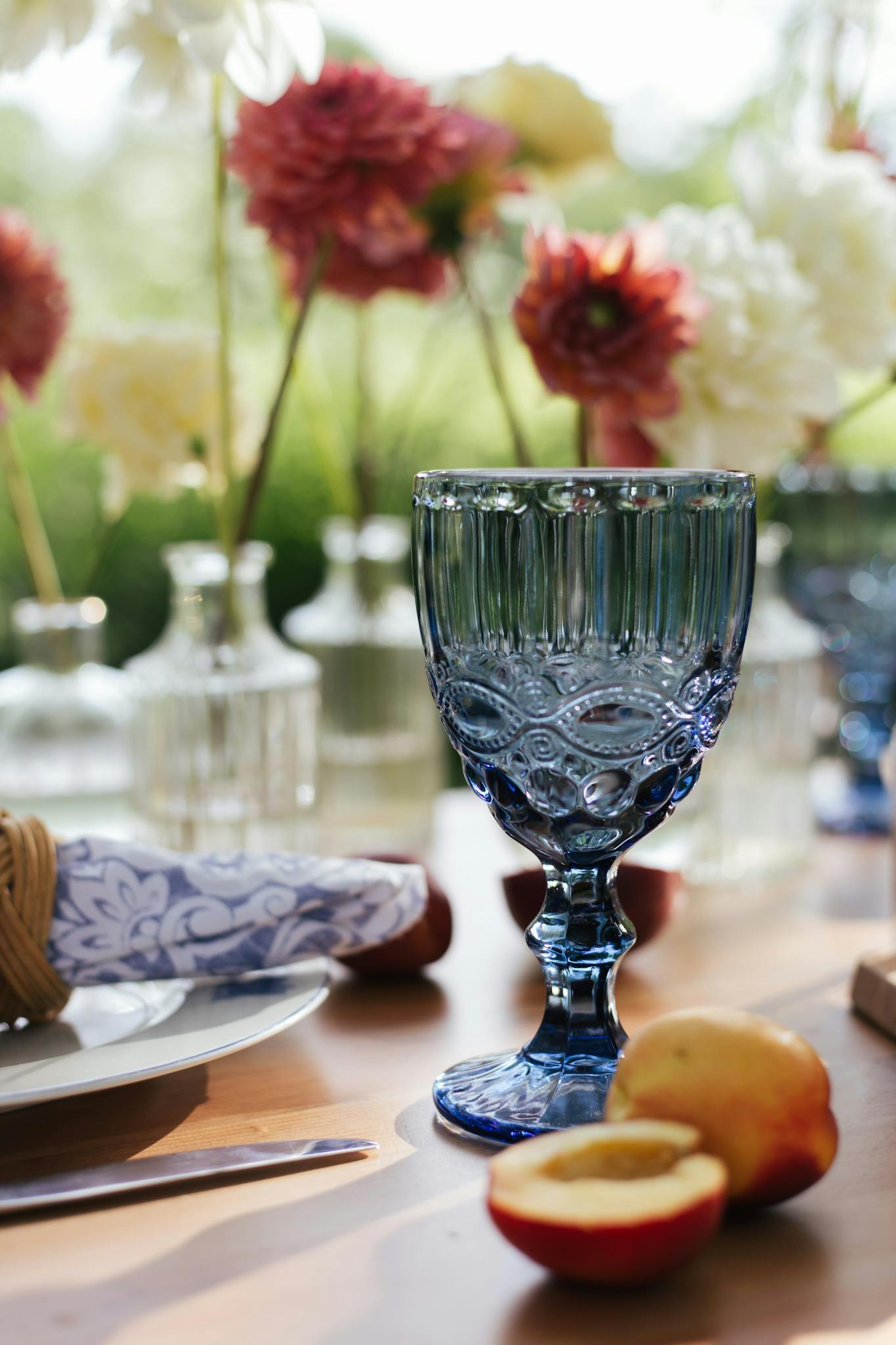 Chic outdoor table scene with blue glass surrounded by flowers and fruit.