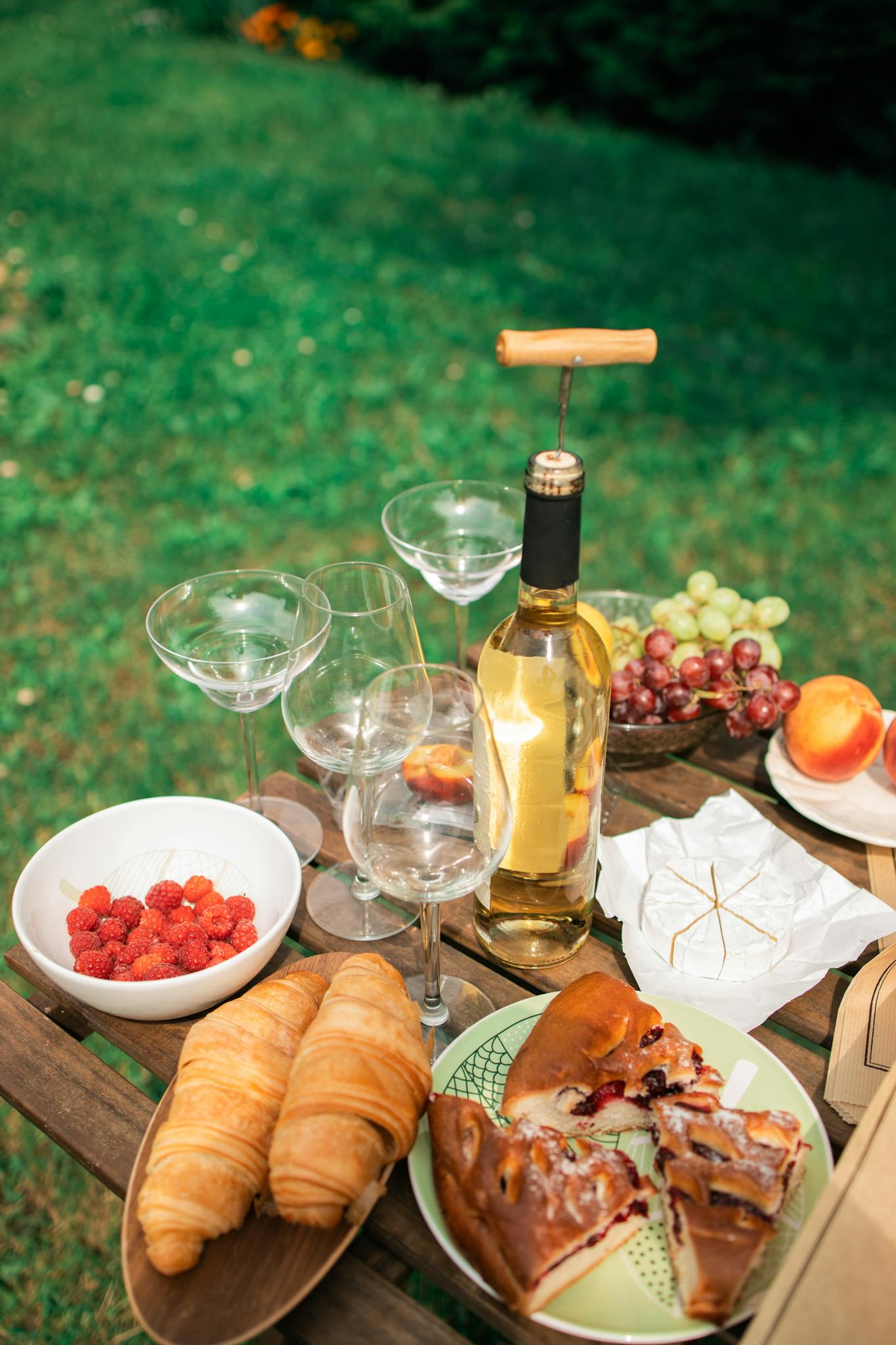 Outdoor picnic setup with croissants, fruit, brie cheese, and white wine on a sunny day.