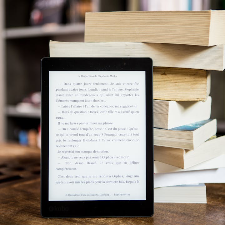 E-reader with text displayed next to a stack of physical books on a wooden table.